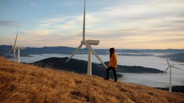 Epic shot of a woman hiking on the edge of the mountain against landscape with wind turbine power station on background. Concept of: environmental engineering, renewable energy and love for nature