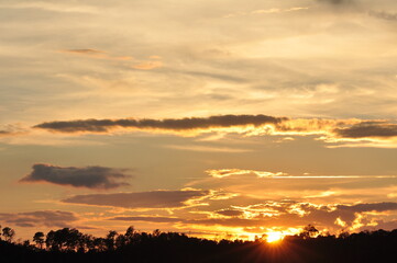 Picture of a sun setting behind a dense forest area followed by mountains.	
