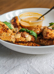 Breaded chicken fingers in a bowl with sauce over wooden table and kitchen towel. Healthy appetiser. Side view. 