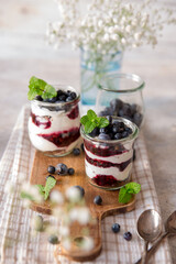 Dessert di yogurt e confettura di mirtilli a strati decorati con menta e mirtilli freschi con fiori bianchi sfocati di fronte e vasetto blu di fiori dietro soggetti