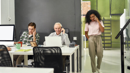 Aged man, senior intern talking on the phone while using laptop, sitting at desk, working together with other employees in modern office