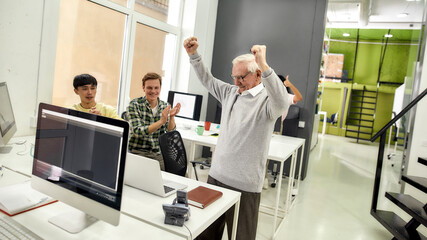 Aged man, senior intern looking cheerful, raising his arms after completing first task at work, Friendly workers applauding, cheering new employee in the office