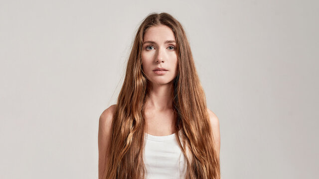 Portrait of young caucasian woman with long hair wearing white shirt looking at camera while standing isolated over grey background