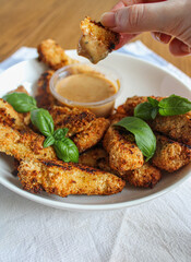 Hand holding chicken finger and dipping into the sauce. A plate of crunchy oven-baked chicken fingers with honey mustard sauce. 