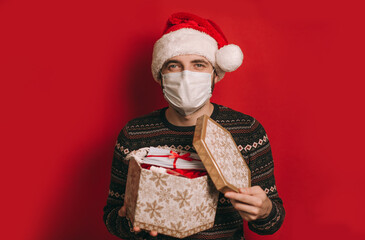 A young man in a medical mask and a red Christmas hat with a gift in his hands, a package of white medical masks and sanitizer. Coronavirus pandemic, quarantine, 2020, 2021. Celebrating the new year