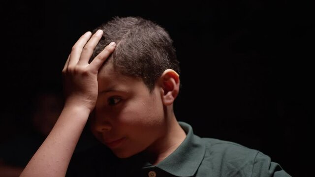 Young hispanic boy sitting at desk, he puts his hand to his head room goes dark