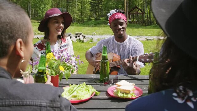 Medium closeup of young joyful friends chilling outdoors in summer time playing guitar and singing enjoying company and nature