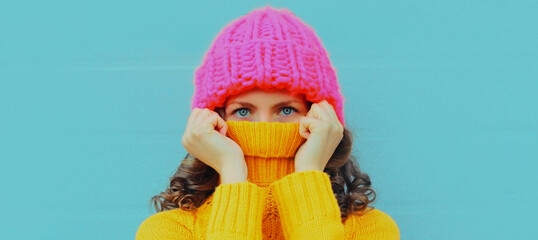 Winter portrait of cute young woman pulls a hat over her face wearing colorful yellow knitted sweater and pink hat on blue background