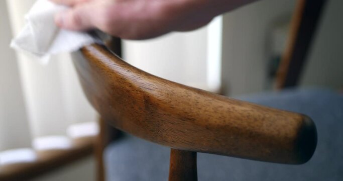 A hand cleaning dust and germs off of wood chair furniture with an antibacterial sanitizing disinfecting wipe to keep the family home healthy and safe from corona virus or bacteria.