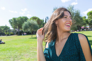 Outdoor, day, in a park portrait of smiling european woman with long hair and dark green dress with hands on head in her hair