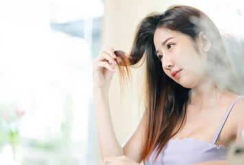 Beautiful young Asian woman holding her long hair with looks at damaged dry hairs,split ends of hair care problems while waiting coffee at cafe. Hair Damage,Haircare, Health And Beauty Concept.