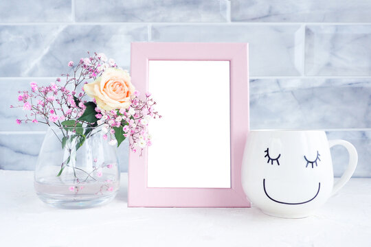 mockup photo frame, white mug with a smiling face and closed eyes and a vase with a rose and gypsophila.