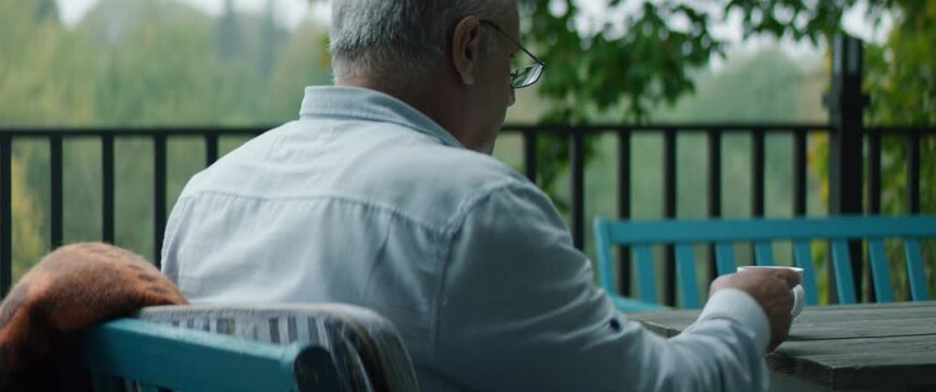 Senior adult mature Caucasian male having a cup of coffee and reading a book on an outdoor terrace of his house on a misty morning. Happy retirement concept