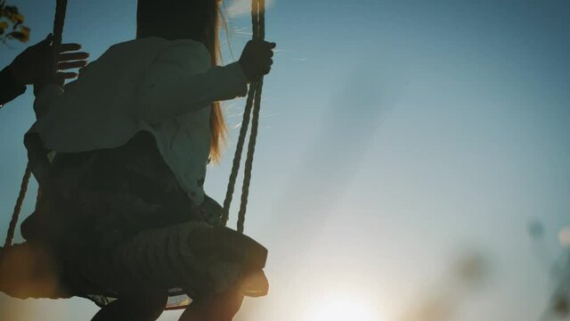Happy young mother and little daughter on a swing at sun light. Pretty girl sitting on a wooden swing and looking at golden sunset. Happy family concept.