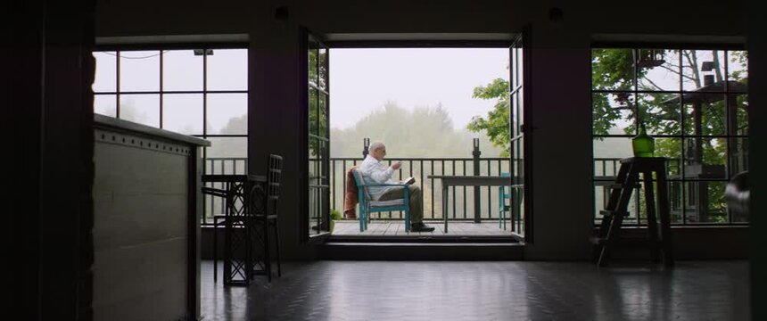 Senior adult mature Caucasian male reading a book on an outdoor terrace of his house on a misty morning. Happy retirement concept. Shot on RED cinema camera with 2x Anamorphic lens
