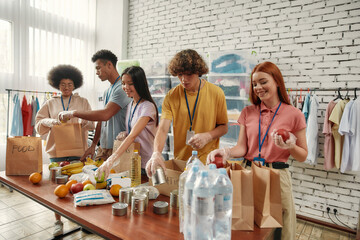 Young male and female volunteers packing food and drinks donation for homeless into paper bags,...