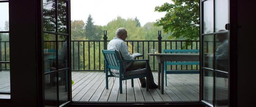 Senior adult mature Caucasian male having a cup of coffee and reading a book on an outdoor terrace of his house on a misty morning. Happy retirement concept