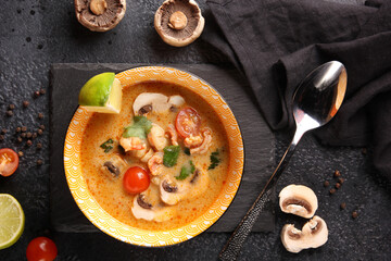 Oriental cuisine, Thai. Tom Yam soup with prawns, shrimps, mushrooms, cherry tomatoes and lime and coriander in a deep bowl and spoon on a black table. Restaurant menu. Background image, copy space