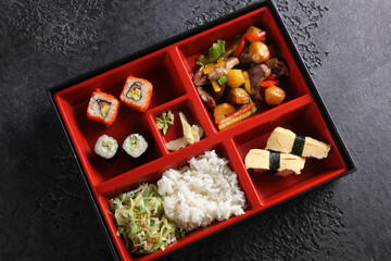 Japanese cuisine. Business lunch in a box: rolls, sushi, rice with vegetables and stew on a black table