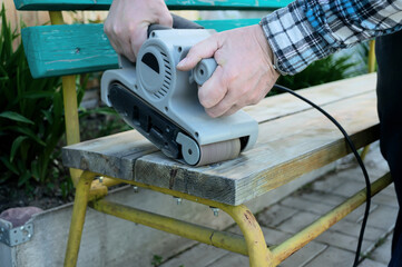Landscaping of the garden. handyman sharpens a wooden bench on the street in the garden. master removes a layer of dry paint from the wood surface using an electric sander.