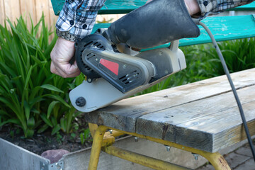 Fototapeta premium Landscaping of the garden. handyman sharpens a wooden bench on the street in the garden. master removes a layer of dry paint from the wood surface using an electric sander