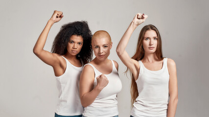 Portrait of three young diverse women wearing white shirts having confident look while showing, raising clenched fist, posing together isolated over grey background - Powered by Adobe