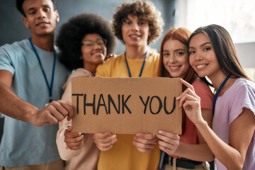 Group of diverse young volunteers smiling at camera, holding card with Thank you lettering while...