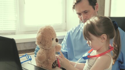 Caring professional male pediatrician playing with a small child in the office. little girl plays with a teddy bear toy listens to him with a stethoscope. toddler patient sits on lap of pediatrician.