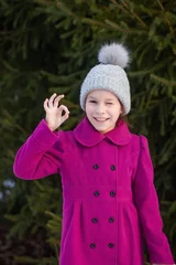 Foto op Aluminium Glimlachend mooi jong meisje in roze jas toont ok, tegen groen van winterpark. © Albert Ziganshin