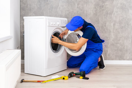 Plumber in overalls with tools is repairing a washing machine in the house