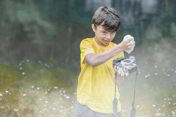 A boy in a yellow t-shirt and shorts is standing near the water in haze outdoor and washing a...