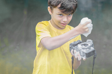 A boy in a yellow t-shirt and shorts is standing near the water in haze outdoor and washing a...