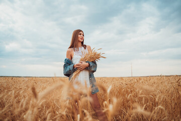 A beautiful young woman in a white dress and jeans stands in a wheat field.