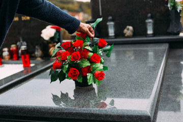 Old woman laying down bouquet of artificial flower on a grave - obrazy, fototapety, plakaty