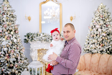 A young man holds a baby in his arms. Father plays with a girl in a white dress, hugs her, kisses against the background of festive Christmas trees, a white fireplace, gifts. Copy space, happy family
