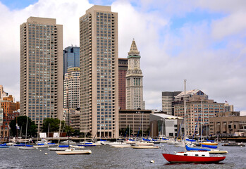Fototapeta na wymiar Scenic cityscape of Boston skyline and waterfront with tall modern buildings, sailboats anchored in harbor, and the Custom House Tower (designated a Boston Landmark).