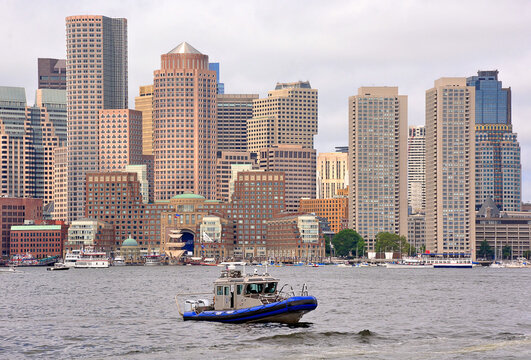 Scenic cityscape of Boston skyline and waterfront with state police boat, tall modern buildings, and boats anchored in harbor.