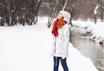 Fototapeta na wymiar Outdoors fashion lifestyle portrait of charming young woman enjoying snowfall in snowy forest. Wearing silver down jacket, red scarf and mittens. Winter river landscape. Enjoying nature and snowfall