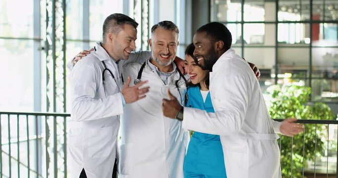 Happy mixed-races males and female doctors standing in clinic, talking and laughing in hugs. Cheerful multi ethnic men and woman physicians communicating, embracing and smiling in hospital.