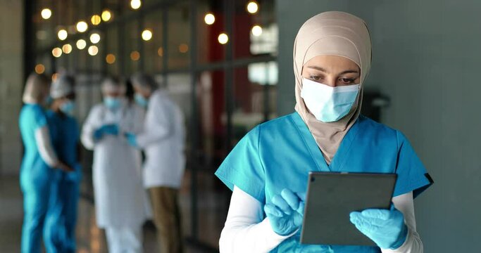 Young Arab woman doctor in hijab, medical mask and gloves standing in hospital and using tablet device. Muslim female medic in traditional headscarf tapping on gadget computer in clinic. Covid-19.