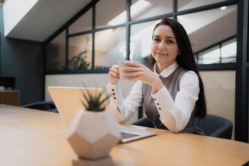 Fototapeta na wymiar a young woman is sitting in a modern office with a Cup of white, hot, fragrant black tea. Looking at the camera, business suit clothes, white shirt