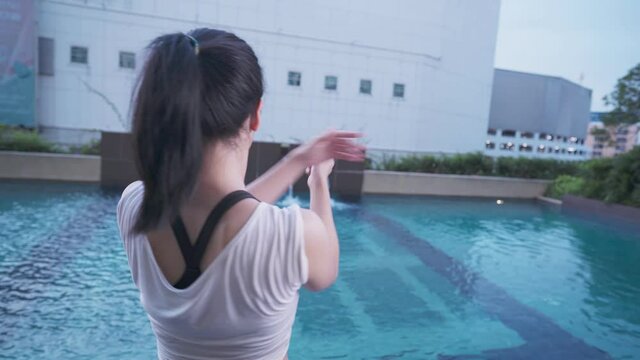 Young Asian woman stretch her arms and shoulder watching swimming pool city high rise resident apartment facility, flexible upper body part, beautiful and relax, exercise motivation, rear view