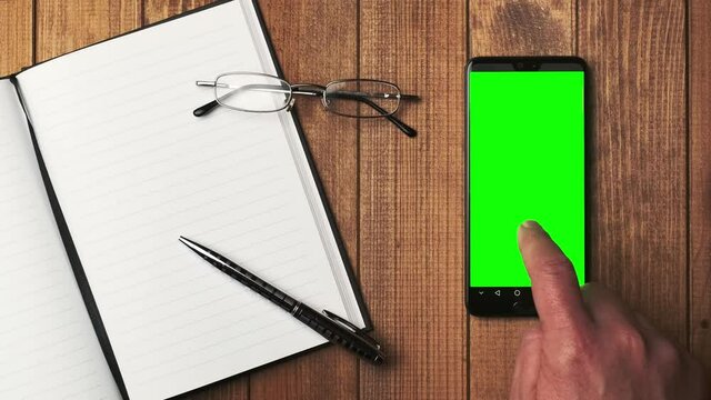 Top view of an open notebook with blank pages, glasses and a cell phone with a green screen on a wooden table. Mobile app mockup.