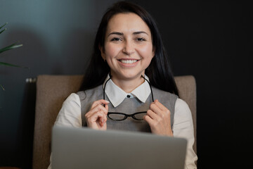 a businesslike young woman looks at the camera and smiles sincerely, holding computer glasses in...