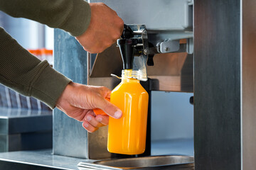 Man fills a plastic bottle with squeezed orange juice from a juicer in supermarket. Fresh orange...