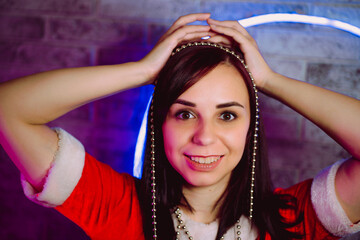 Portrait of young woman in Santa Claus suit with beads against illuminated wall. Close up of pretty female in Christmas costume holding beads. Concept of Christmas celebration.