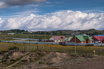 Punta Arenas, Chile - December 12, 2008: Houses in green meadows in rural area outside city center with white belt of huge cloudscape under blue sky.