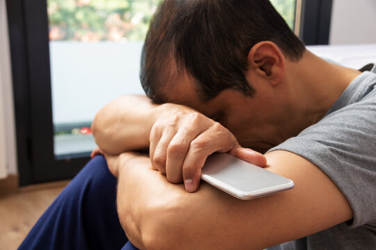 Single sad holding a mobile phone lamenting sitting at the foot of his bed in his bedroom