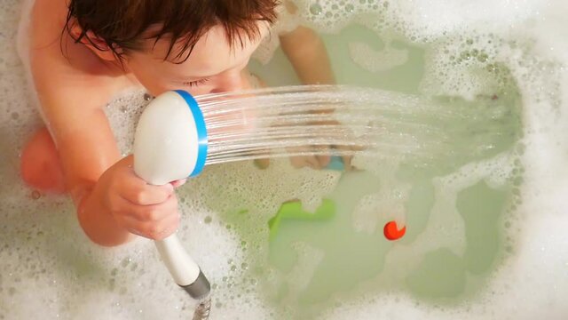 A little boy playing with a shower while sitting in a bubble bath
