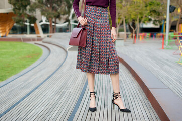 Fashionable young woman wearing pleated midi skirt, sweater, black high heel shoes and holding...